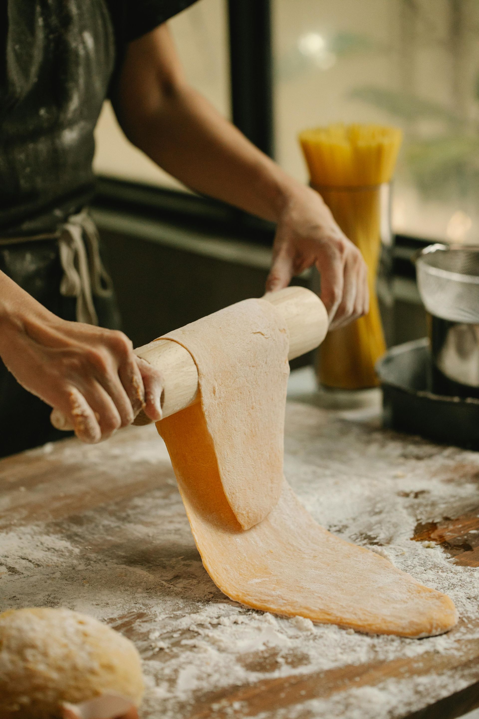 Mani che stendono un impasto arancione su una superficie di legno infarinata. Spaghetti sullo sfondo.