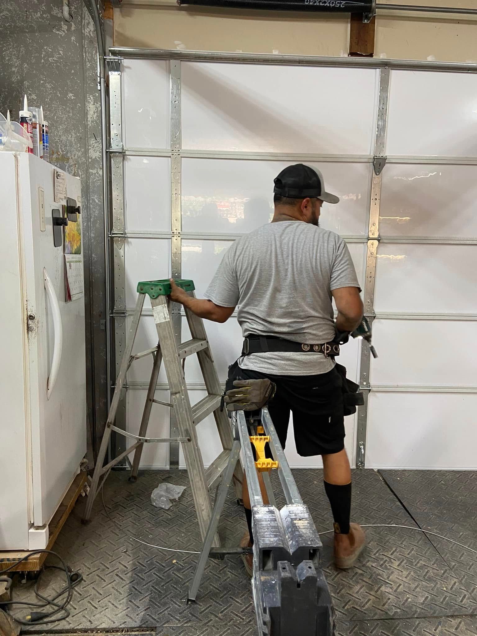 A person in a gray shirt and black shorts stands on a ladder, working on a white metal garage door installation.