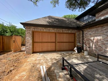 A brick garage with natural wood doors sits beside a fence and a flatbed trailer in a sunny outdoor setting.