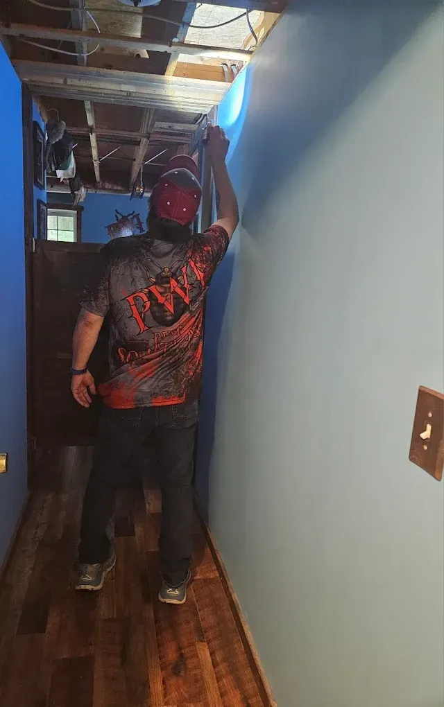 Person standing in a narrow hallway, reaching up toward the ceiling, wearing a red cap and black graphic shirt.