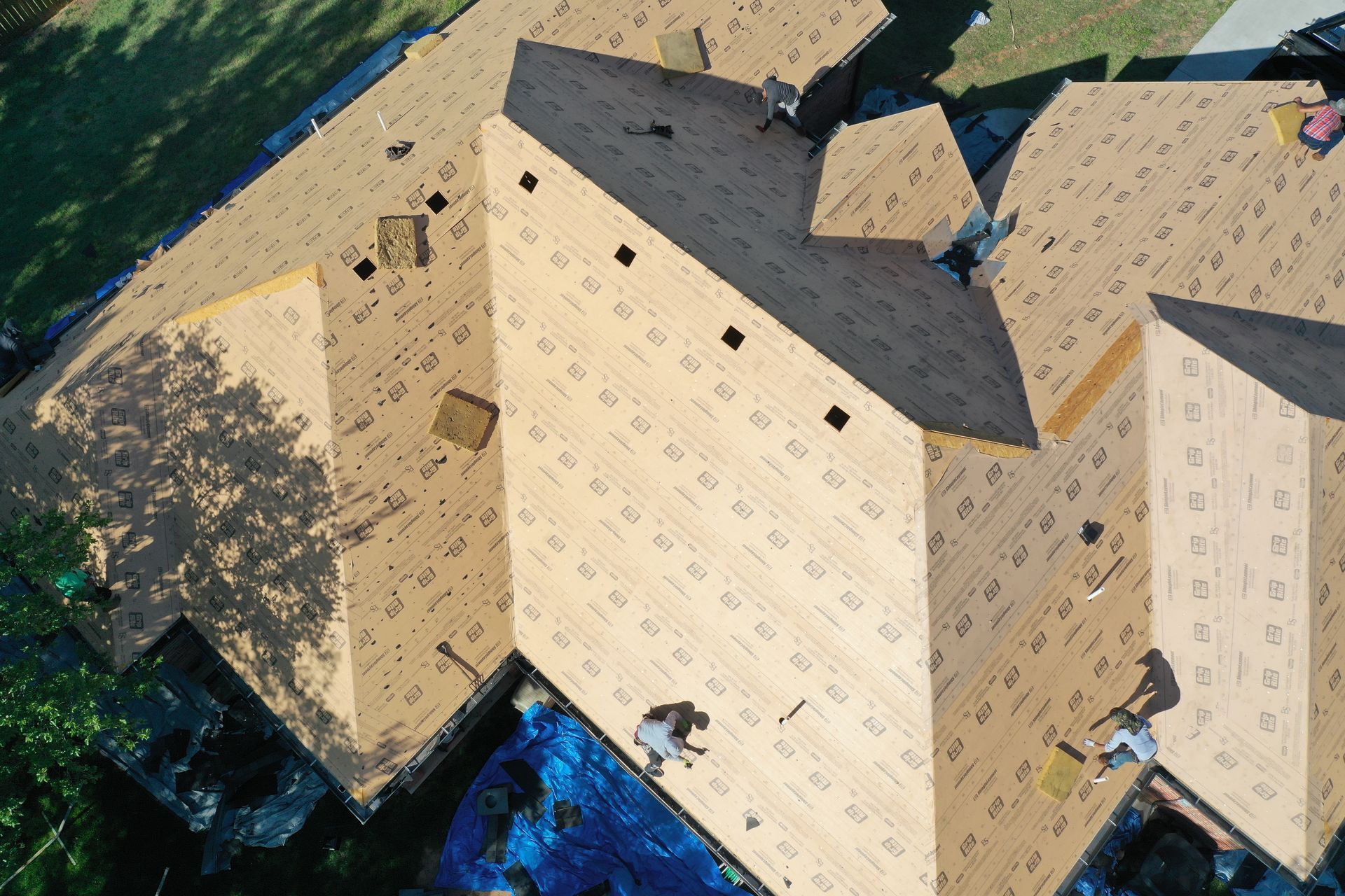 Aerial view of a tan, multi-gabled house roof with blue tarps and surrounding trees.