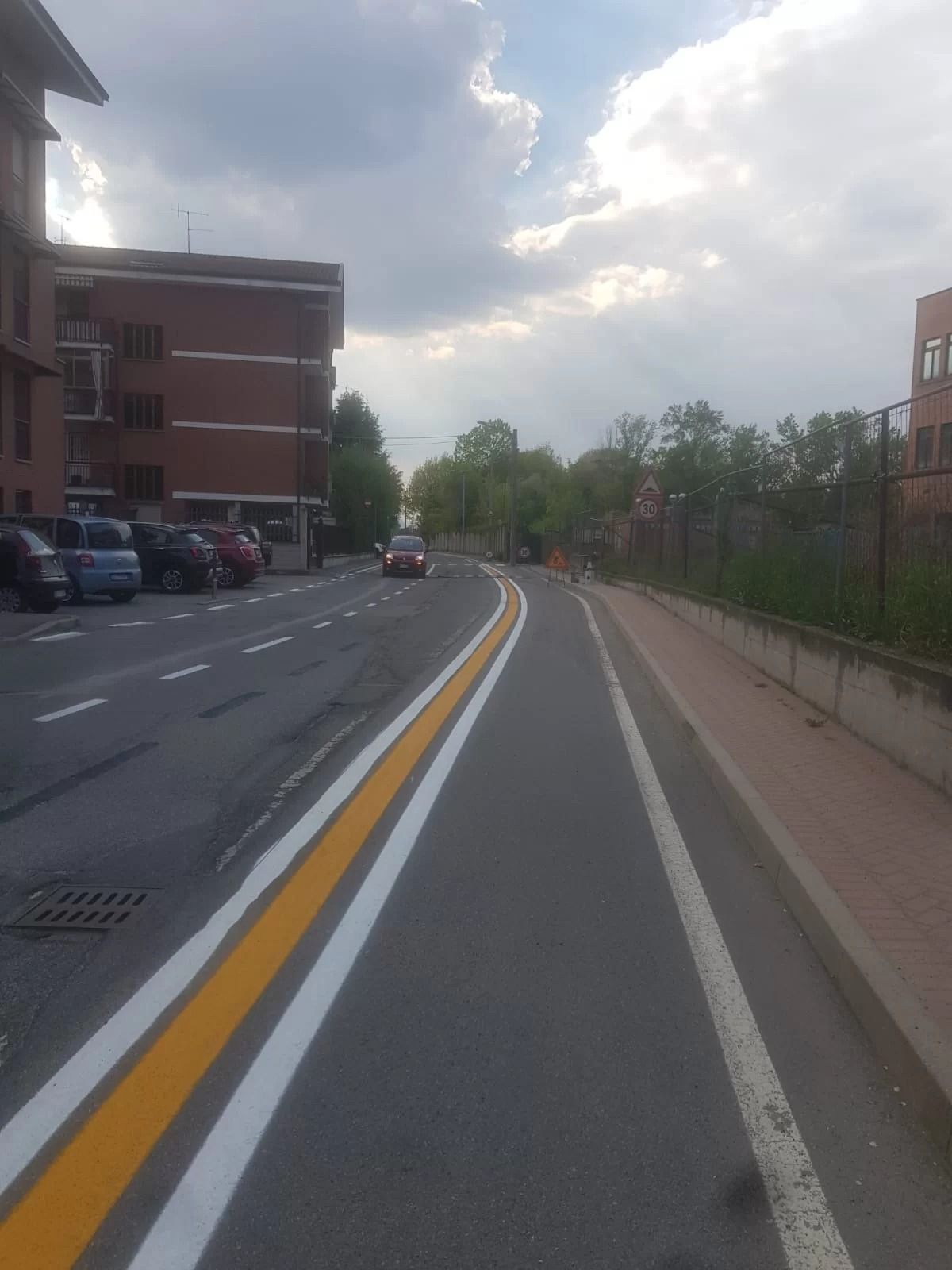 Una scena di strada asfaltata con una pista ciclabile dedicata, segnalata da linee gialle e bianche, delimitata da auto parcheggiate e vegetazione.