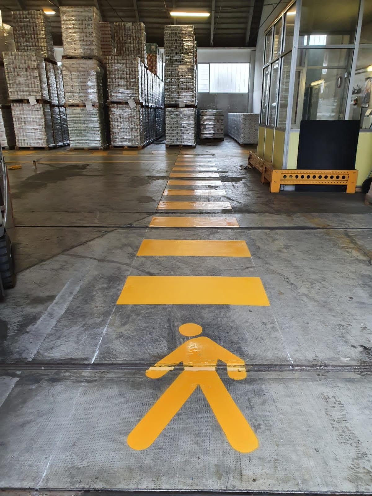 Un attraversamento pedonale dipinto di giallo con l'icona di una persona che cammina, su un pavimento di cemento all'interno di un magazzino con pallet di stoccaggio.