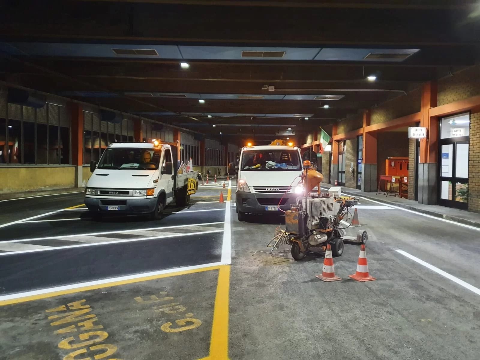 Di notte, alcuni operai utilizzano dei macchinari per dipingere la nuova segnaletica orizzontale bianca su una strada, con due furgoni di servizio parcheggiati nelle vicinanze.