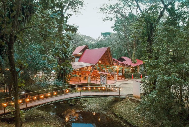Una casa con techo rojo está rodeada de árboles y un puente sobre un río.