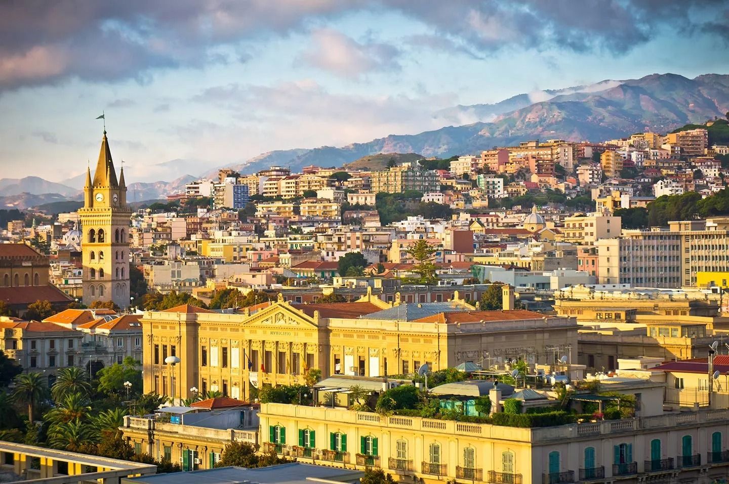 Paesaggio urbano con edifici, una torre dell'orologio e uno sfondo di montagne, il tutto immerso nella luce dorata del sole.