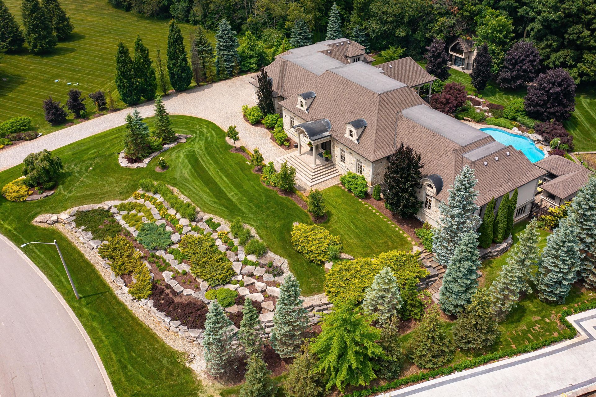 Aerial view of a large estate house with a terraced rock garden, green lawn, manicured trees, and a backyard pool.