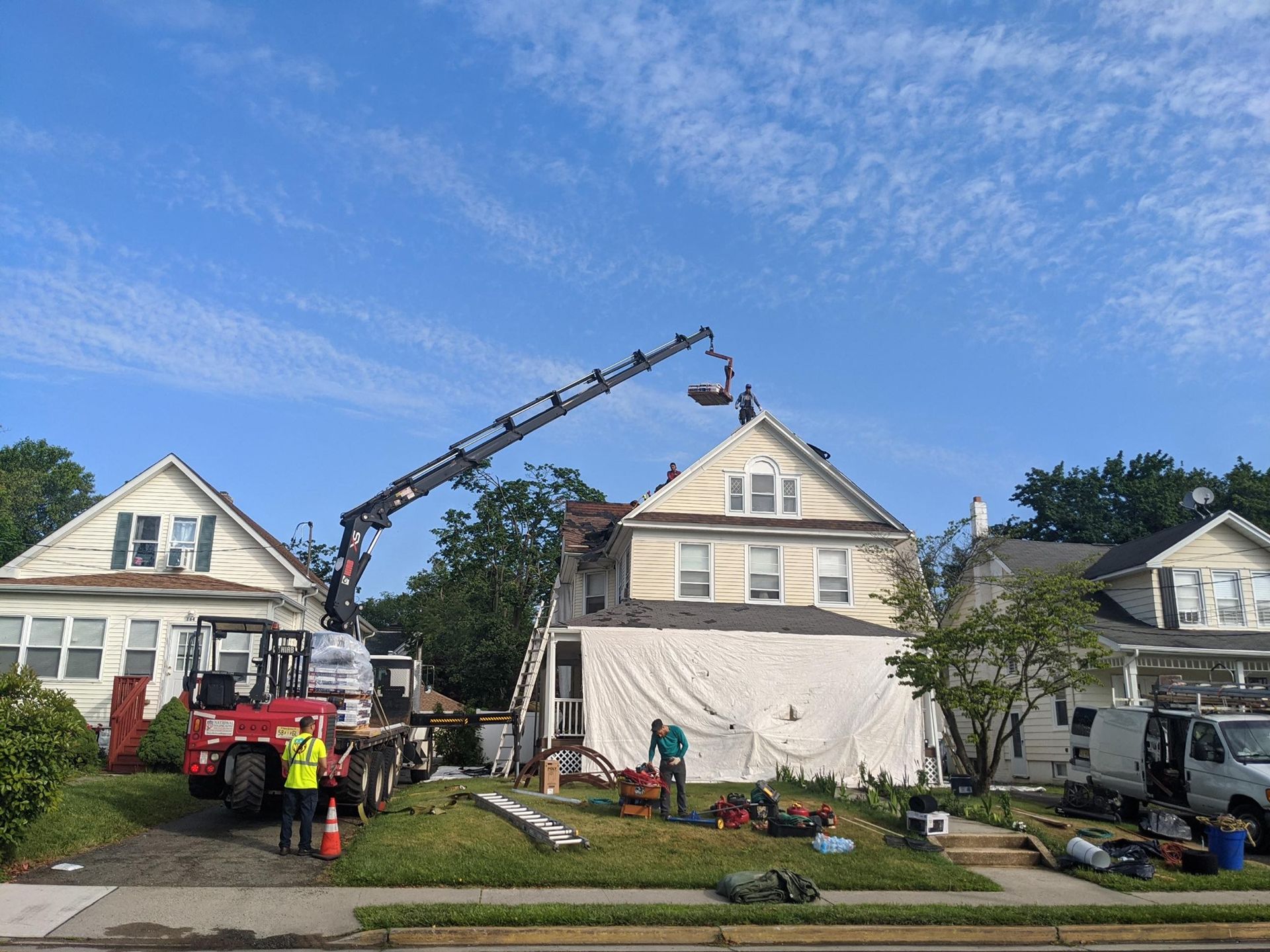 A crane lifts roofing materials to a house roof under construction, with workers and equipment visible.