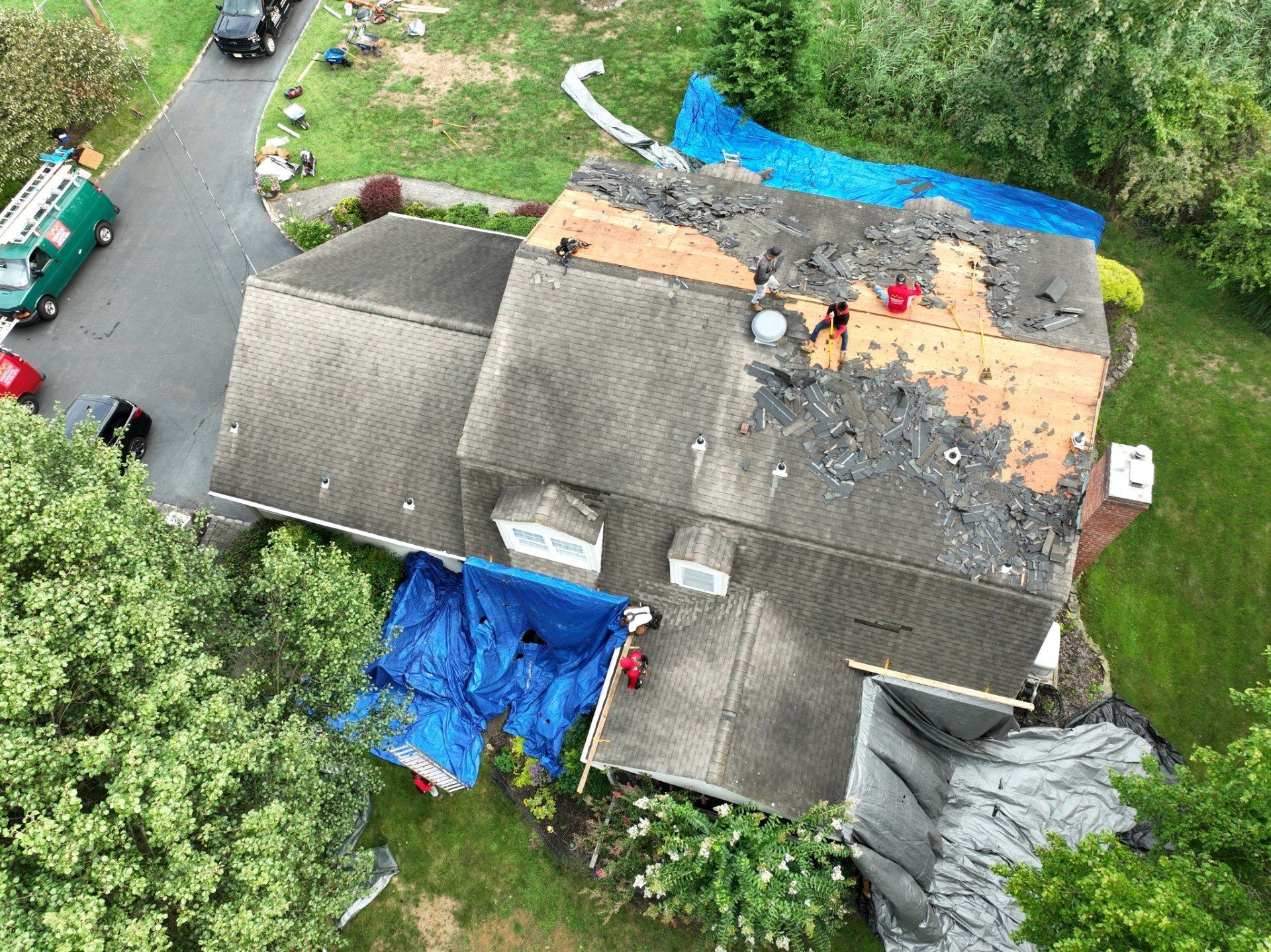 Overhead view of a house roof partially covered by blue tarps, with roofers working. Green truck and surrounding greenery.
