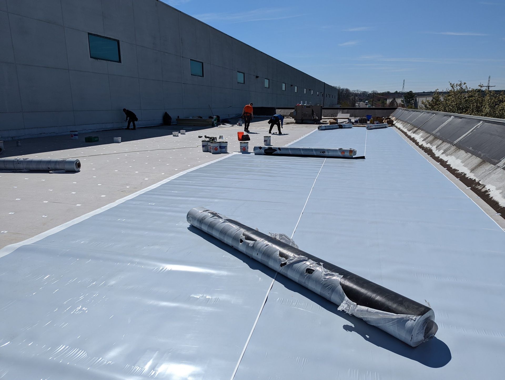Workers installing a light blue roofing membrane on a flat commercial roof under a bright blue sky.