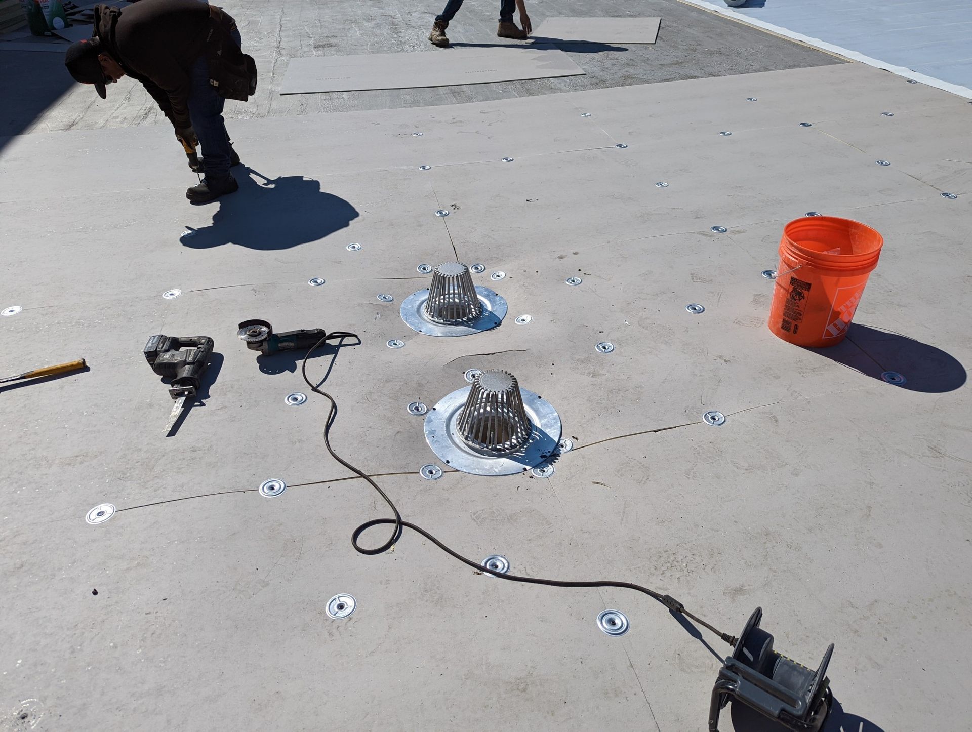 Workers installing rooftop vents on a light-colored surface; tools and a bucket are nearby.