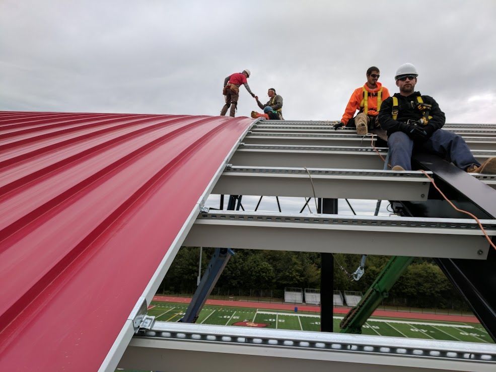 Construction workers installing a red metal roof on a building frame, cloudy sky overhead.