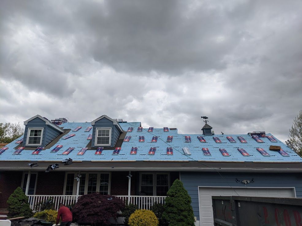 Roof partially covered in blue underlayment, with shingles being installed on a house under a cloudy sky.