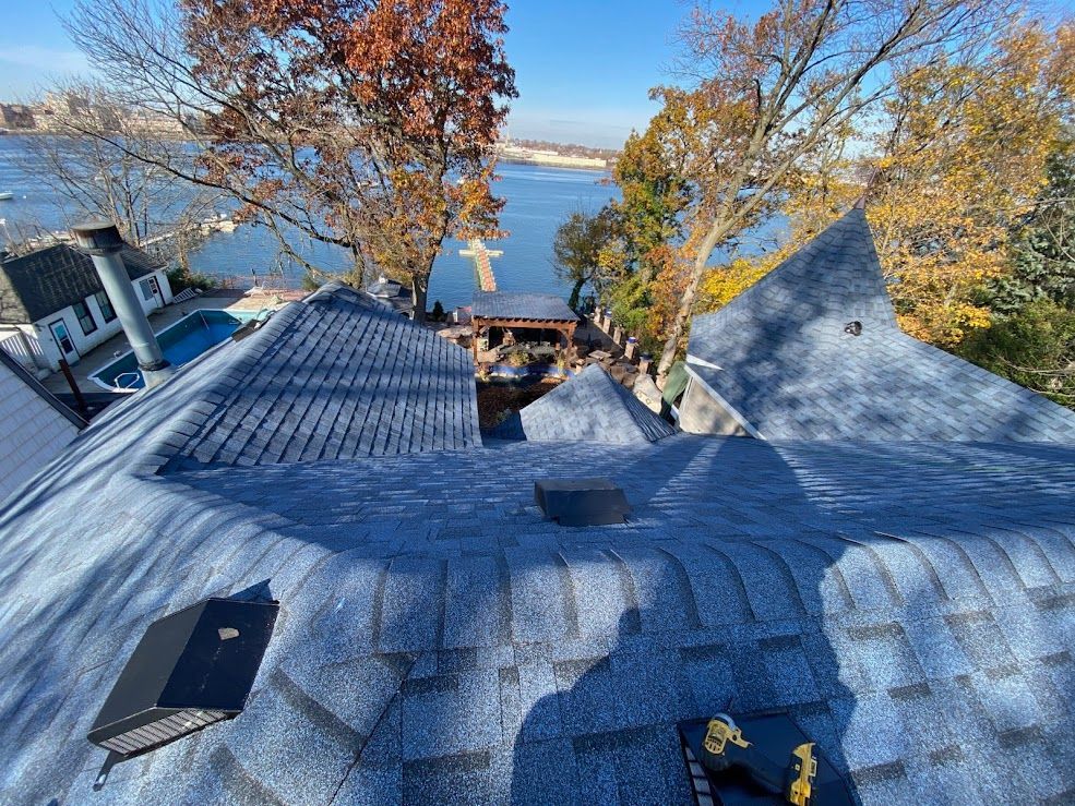 View of a blue-shingled roof with a water and trees background.