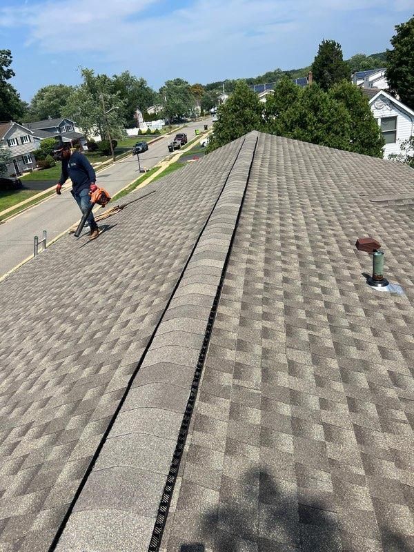 Two workers on a shingled roof repair. A residential street is in the background on a sunny day.