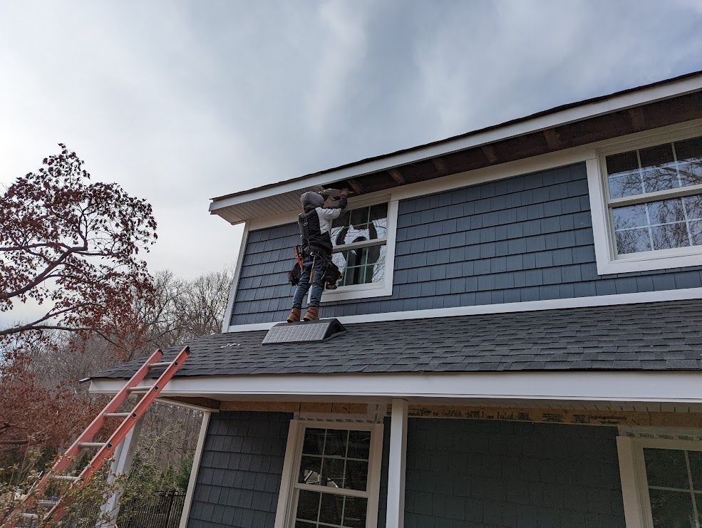 Person on a ladder, installing something on a house's upper side, under cloudy sky.