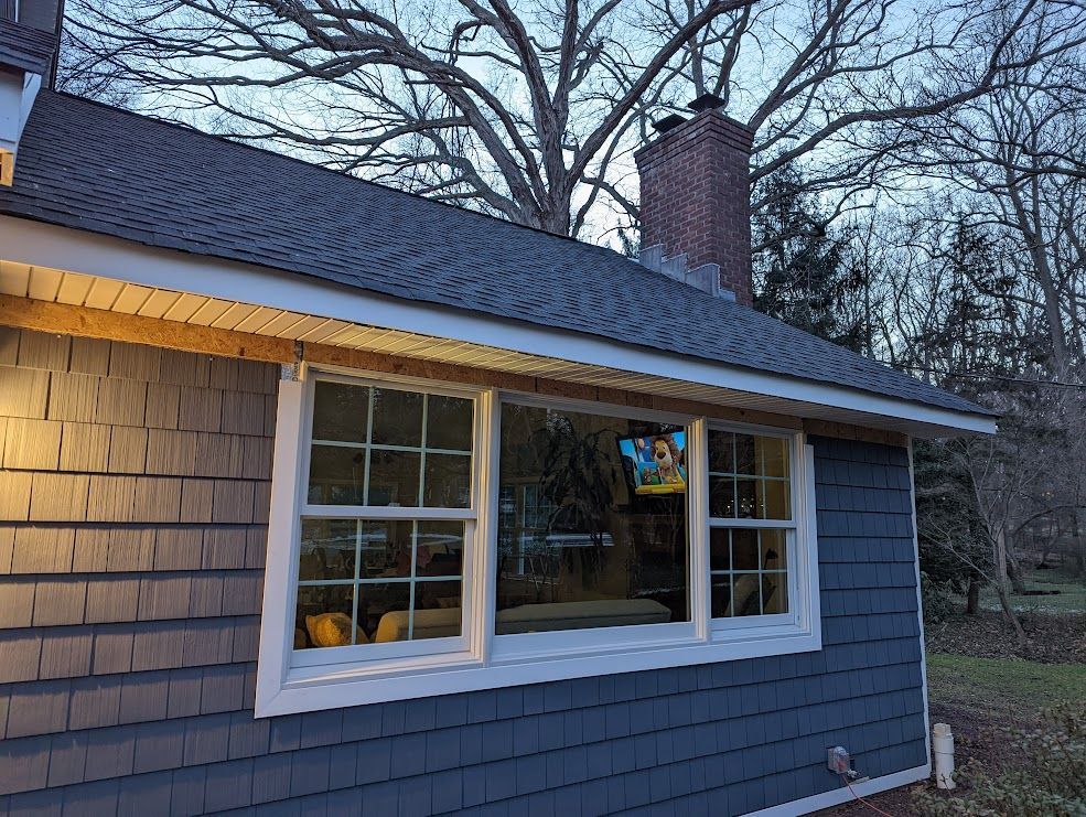 Exterior of a house with gray shingle siding, white trim windows, and a dark roof. A brick chimney rises in the background.
