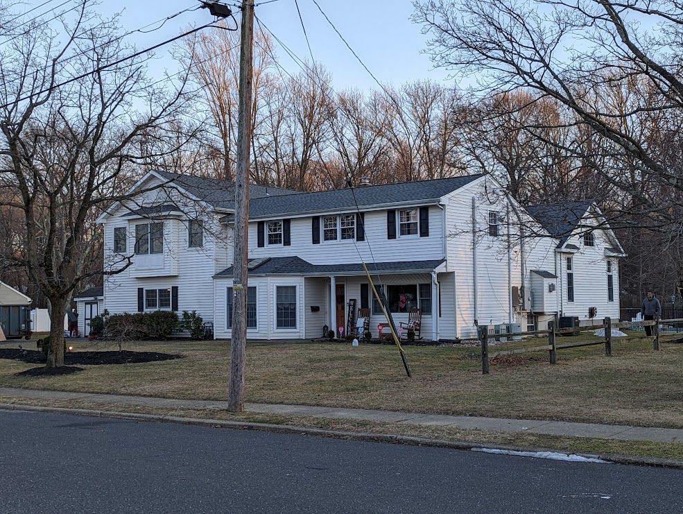 White two-story house with black shutters, trees in the background, and a utility pole in front.
