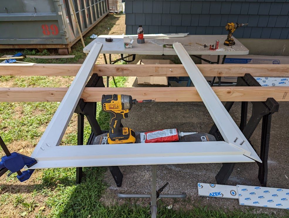 White window frame in a triangle shape on sawhorses, with tools and materials nearby, outdoors.
