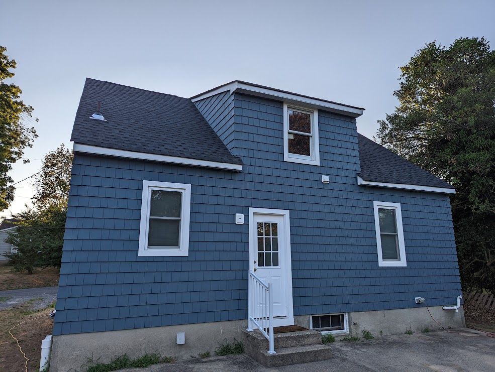 Blue-shingled house with white trim, door, and windows. Dormer with window. Gray roof.