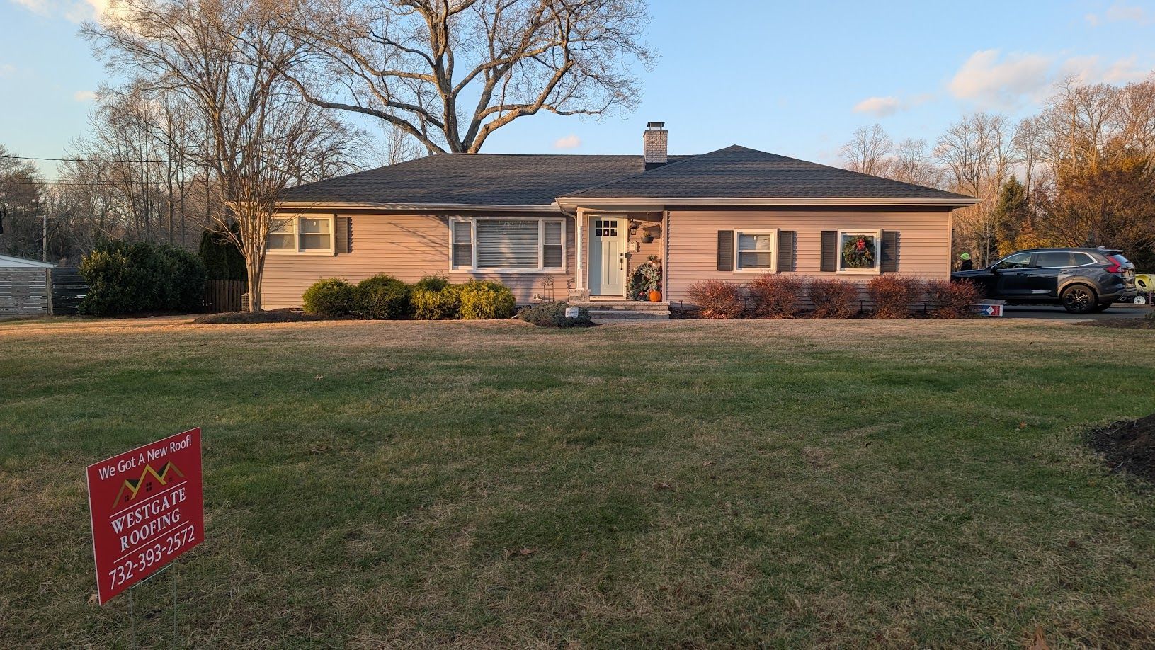 Ranch-style house with pinkish siding, a dark roof, and a lawn with a sign in front. A car is parked on the right.