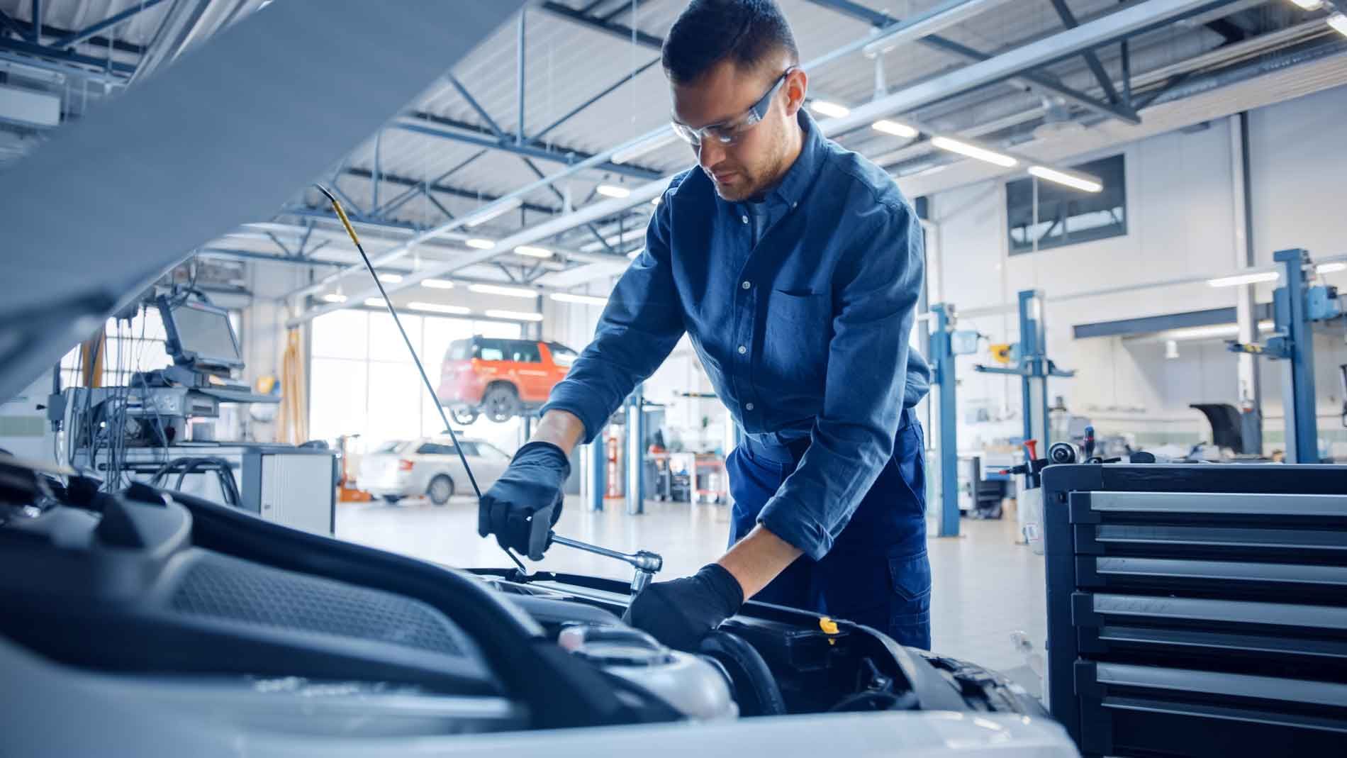 Mekaniker i blå uniform, sikkerhedsbriller, arbejder på bilmotor i et værksted.