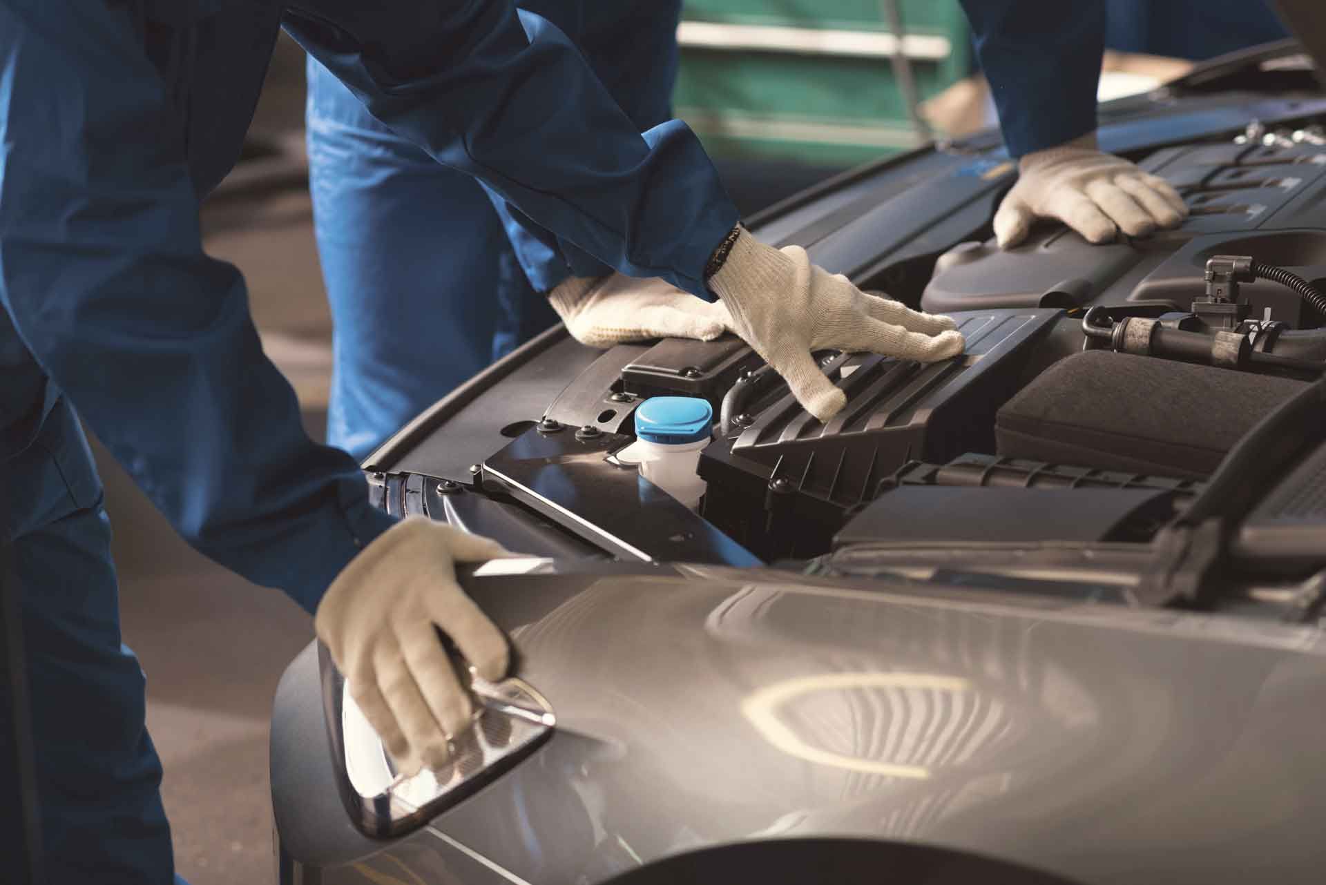 Mekaniker i blå dragt og hvide handsker arbejder på en bilmotor i en garage.