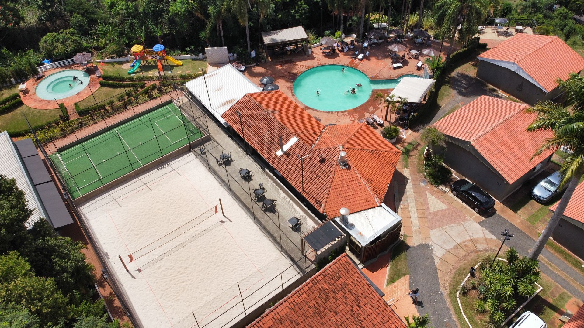 Vista aérea de um resort com piscina, quadra de tênis, quadra de vôlei e telhados de telha vermelha.