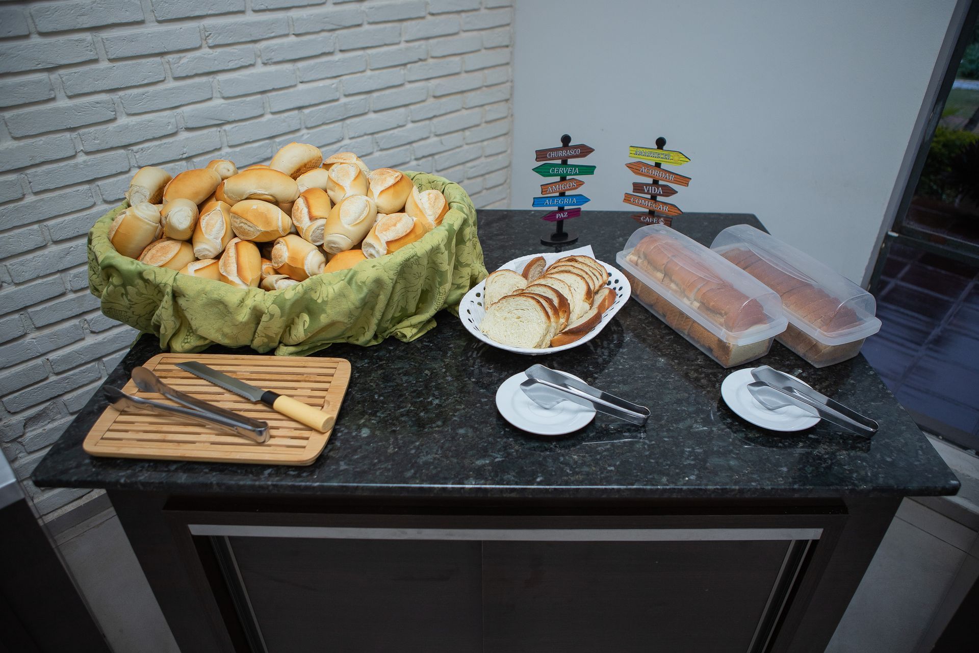 Cesta de pão, pão fatiado, pães de forma e utensílios dispostos sobre uma bancada escura.