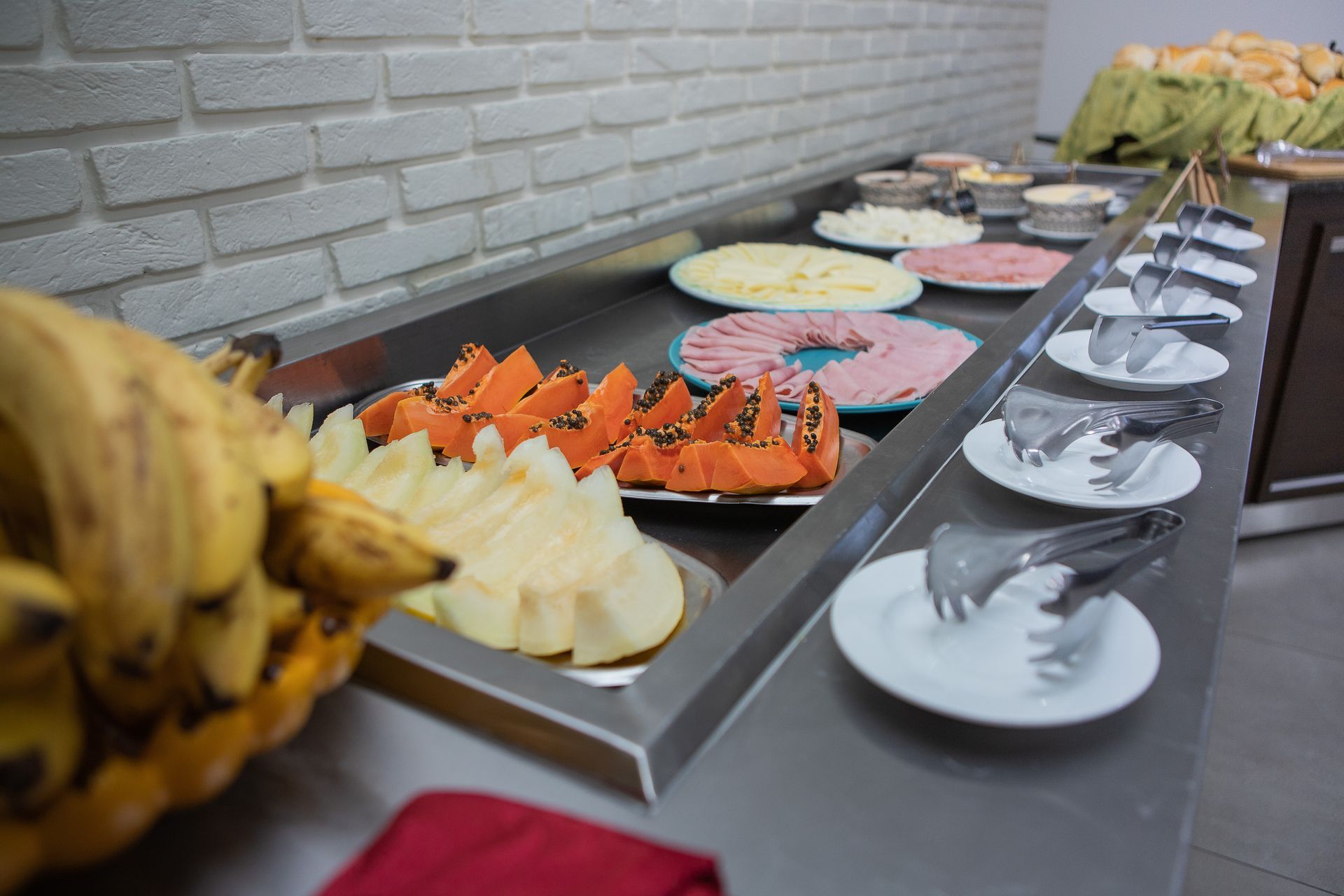 Buffet de café da manhã com frutas, queijo, presunto e utensílios para servir.