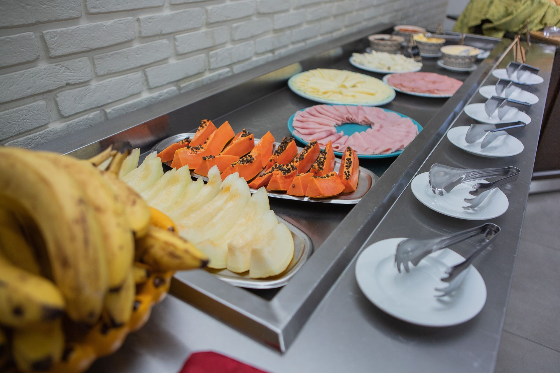 Buffet com frutas fatiadas, presunto, queijo e condimentos, além de pratos e utensílios para servir.
