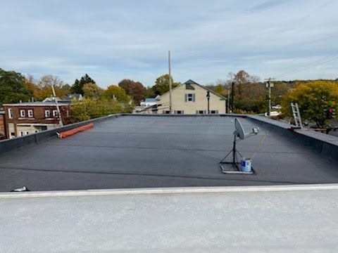Flat black rooftop with a satellite dish, overlooking nearby houses and trees under a cloudy sky