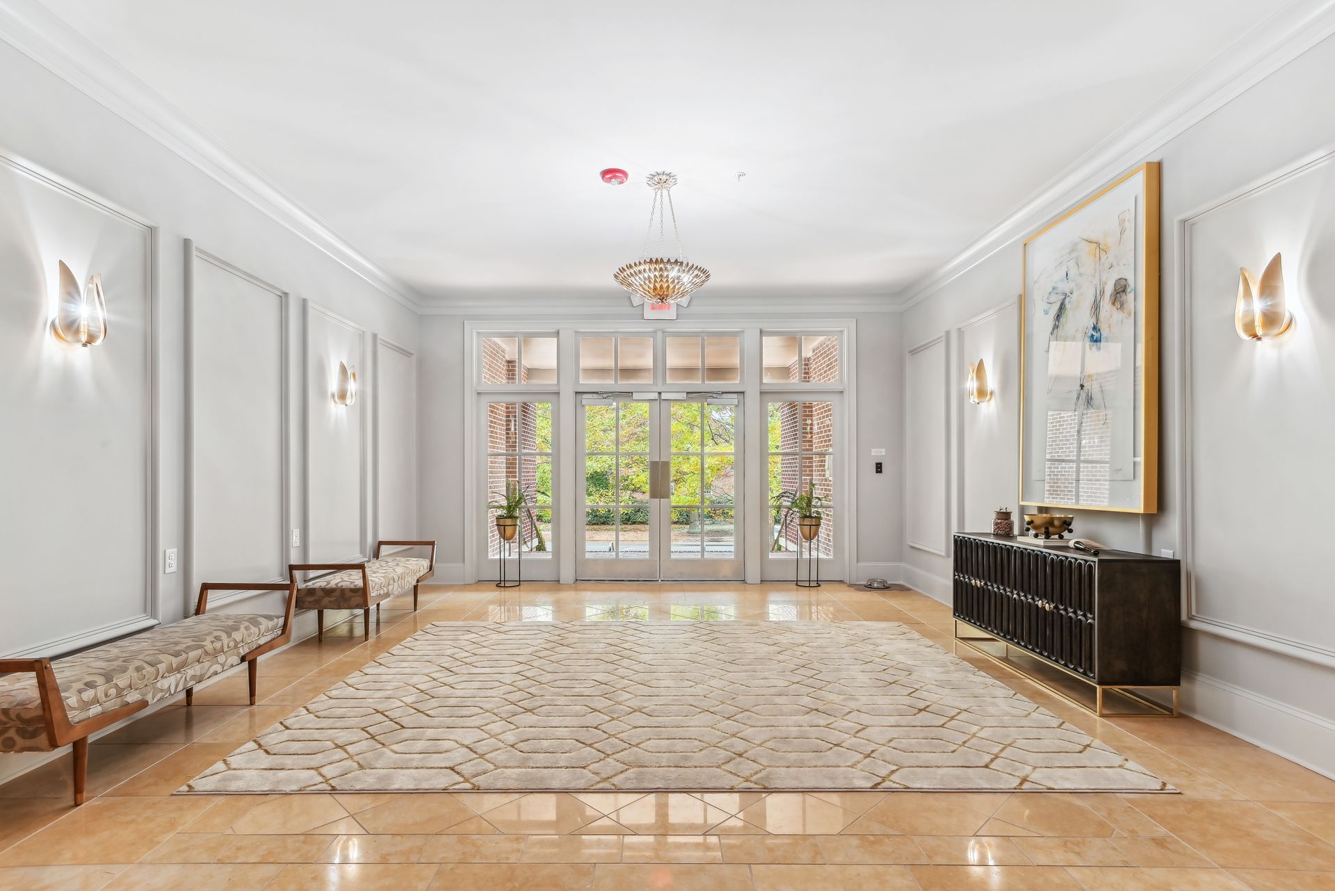 Elegant foyer with light walls, large rug, chandeliers, and outdoor view through double doors.