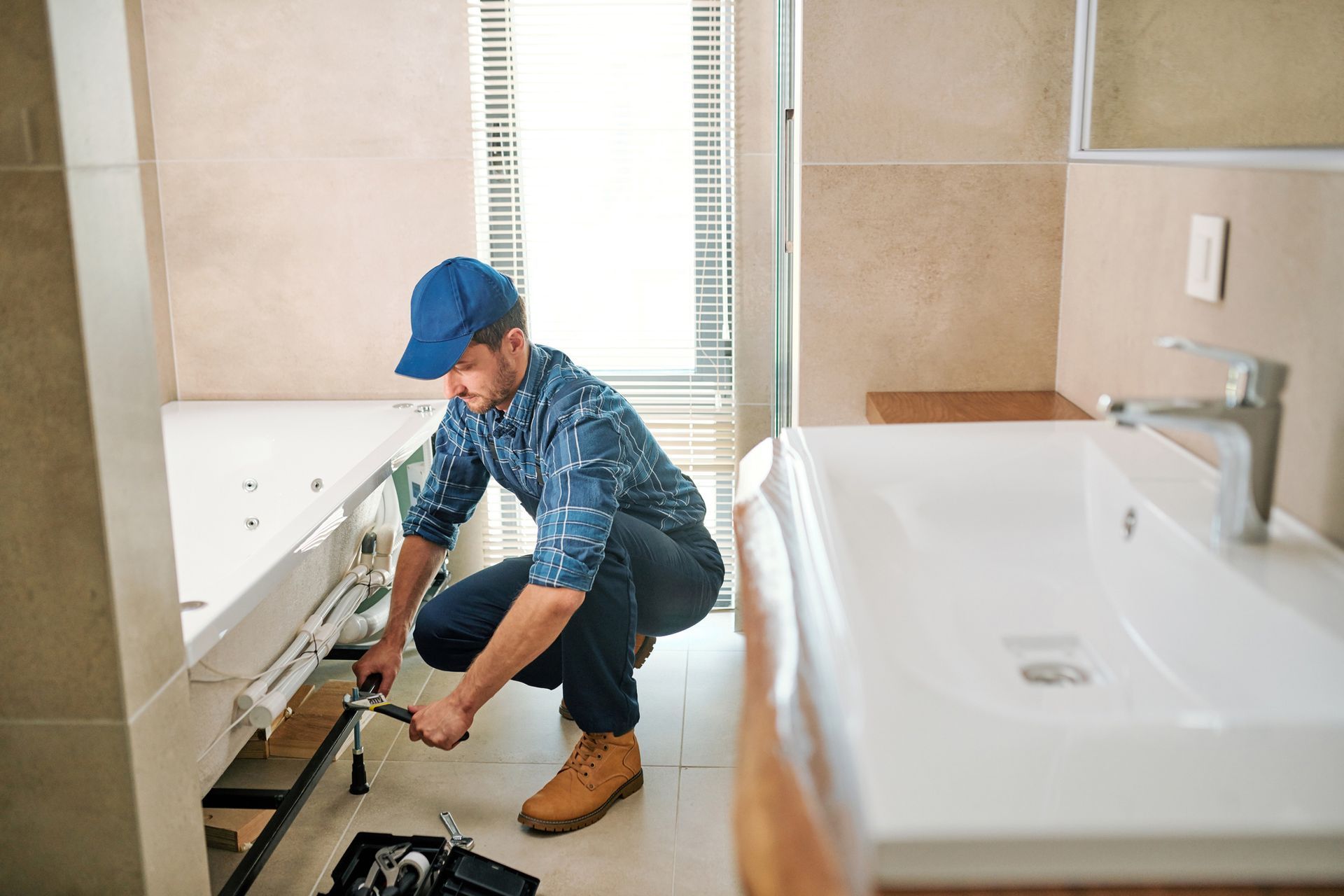 Plumber in blue cap and flannel shirt working on a white bathtub in a bathroom with a window.