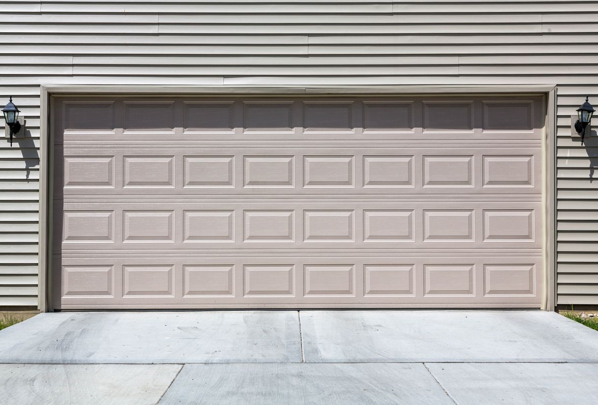 Tan garage door with square panel design; surrounded by light-brown siding and concrete driveway.