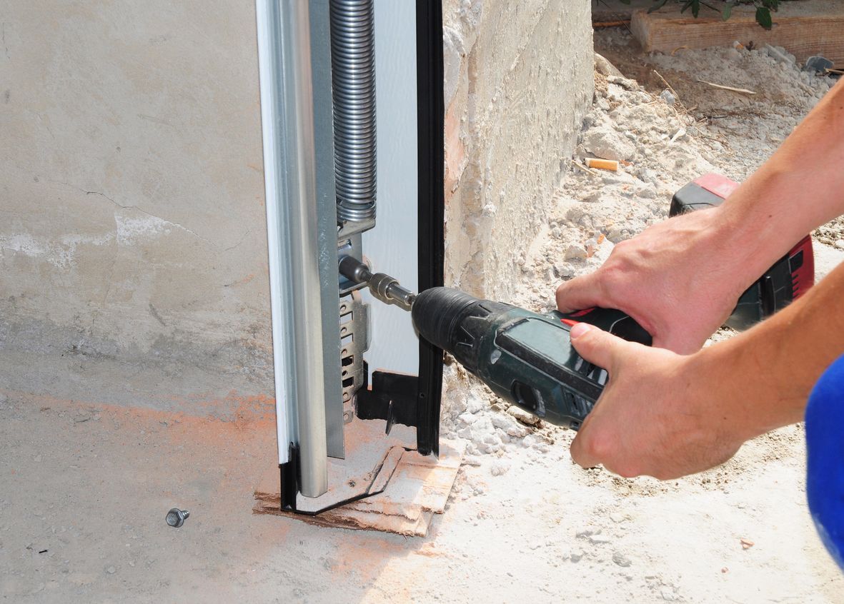 Person using a drill to work on a garage door track; outdoors near a concrete wall.