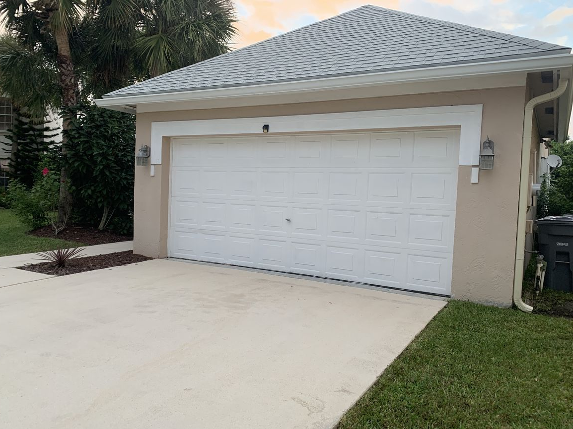 White garage door on a light brown building with a gray roof, next to a driveway and green grass.