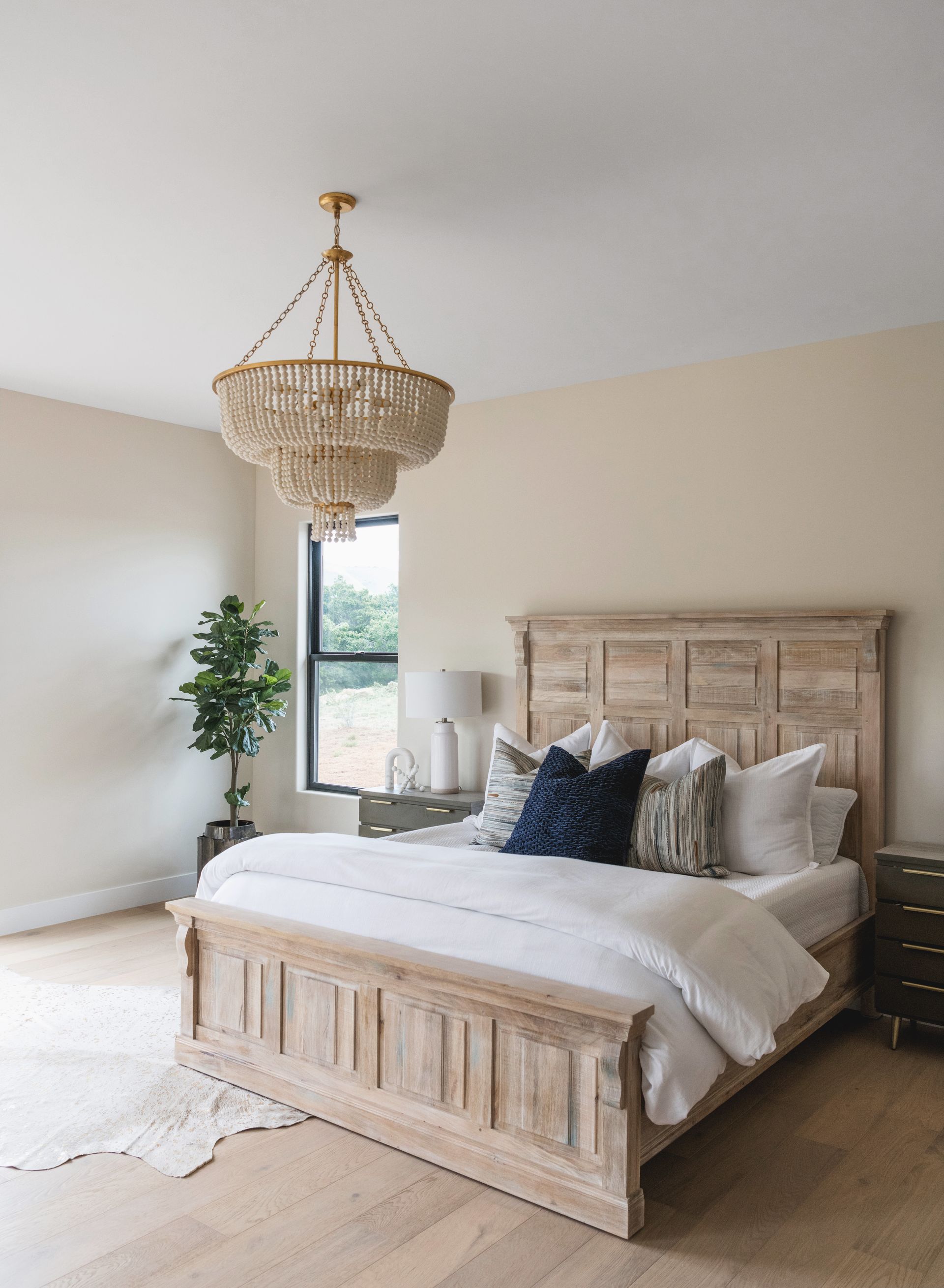 Bright bedroom with a wooden bed, white bedding, woven chandelier, and a potted plant by the window