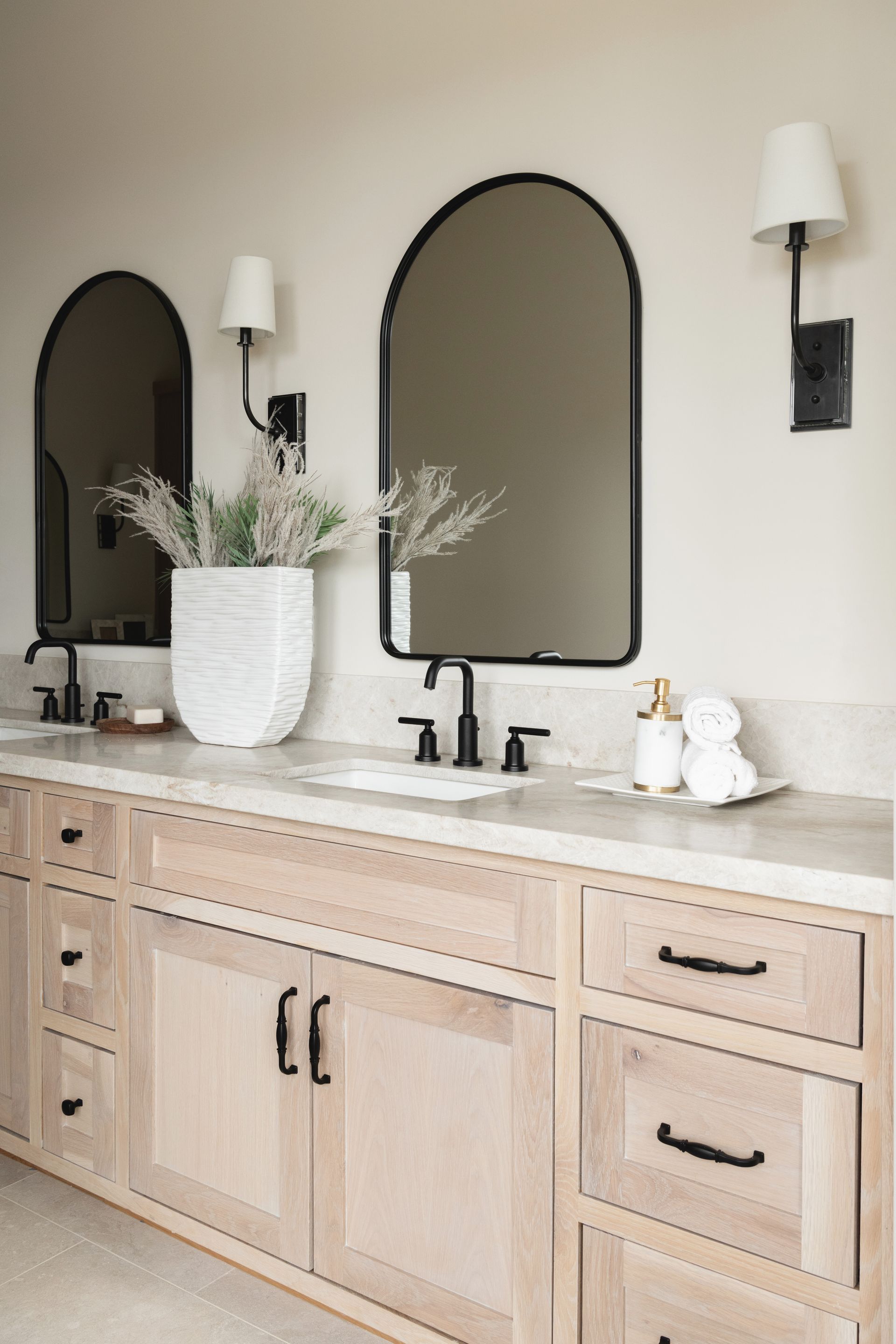 Light wood bathroom vanity with double sinks, arched mirrors, black sconces, and white decor.