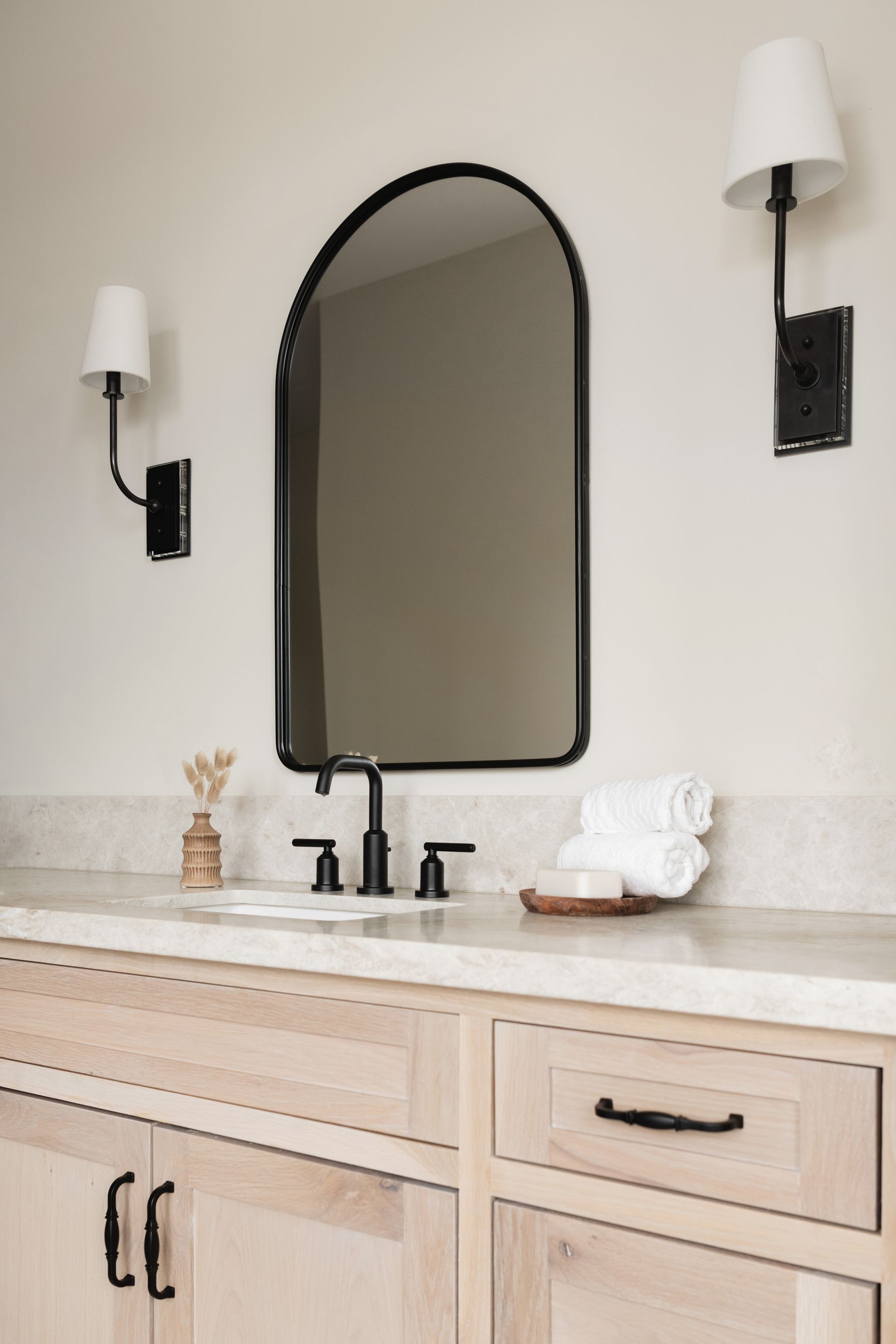 Modern bathroom vanity with arched black-framed mirror, twin wall sconces, and beige cabinetry