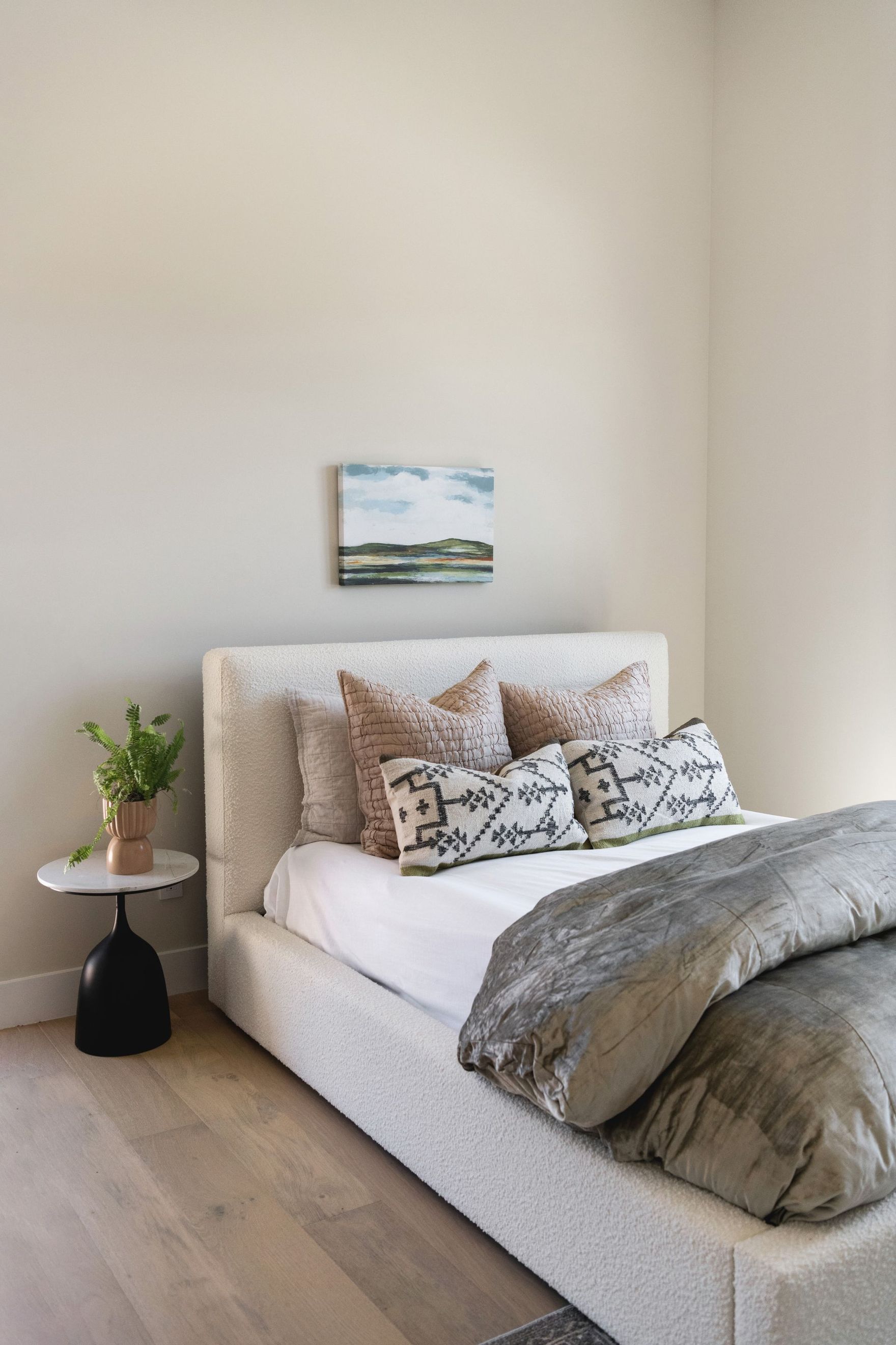 Cozy bedroom with a neatly made bed, gray bedding, patterned pillows, and a small side table with a plant.
