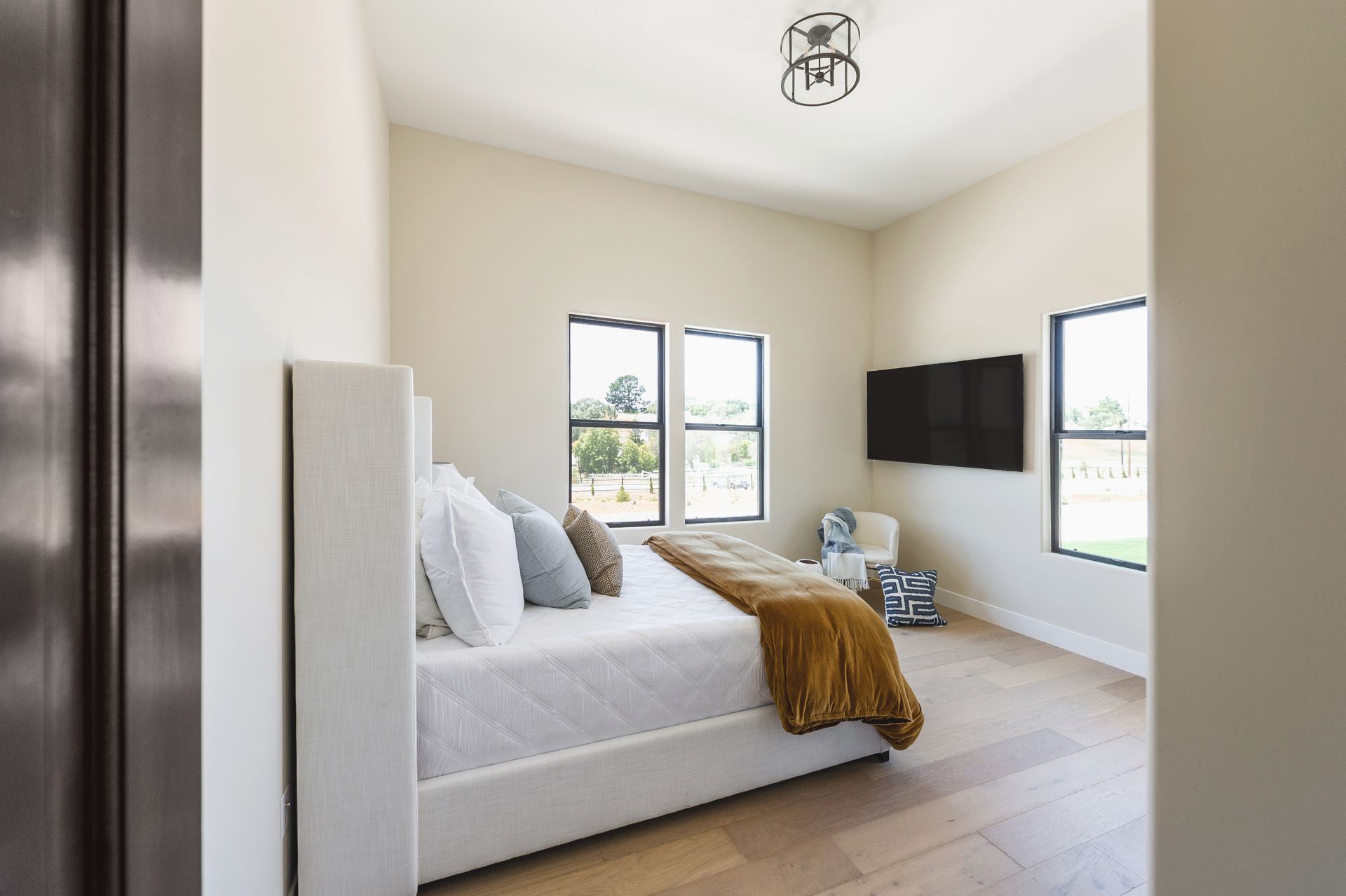 Modern bedroom with a white bed, gray pillows, tan throw, wall-mounted TV, and windows on two sides