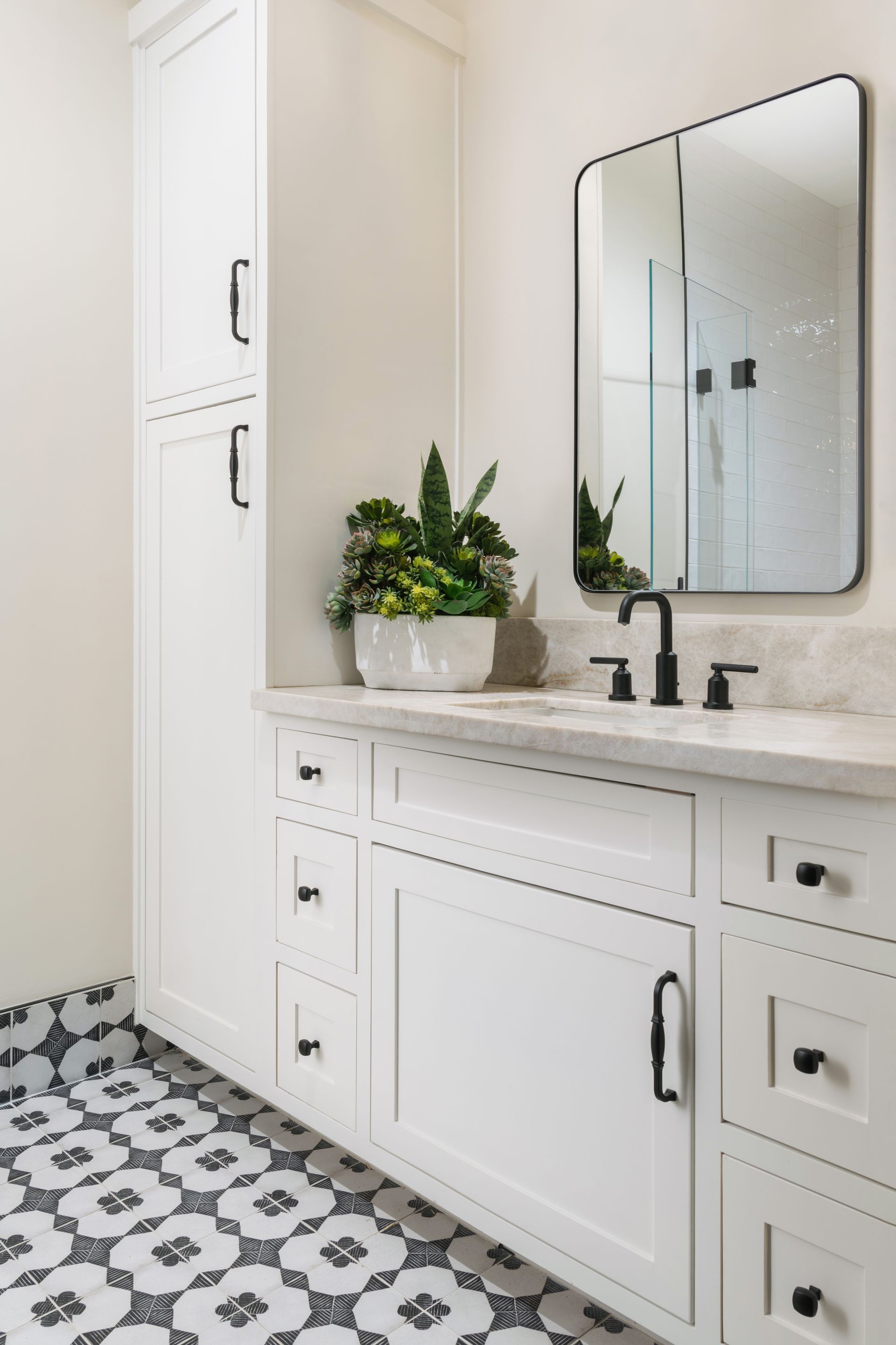 Bright white bathroom vanity with mirror, black faucet, potted plant, and patterned tile floor