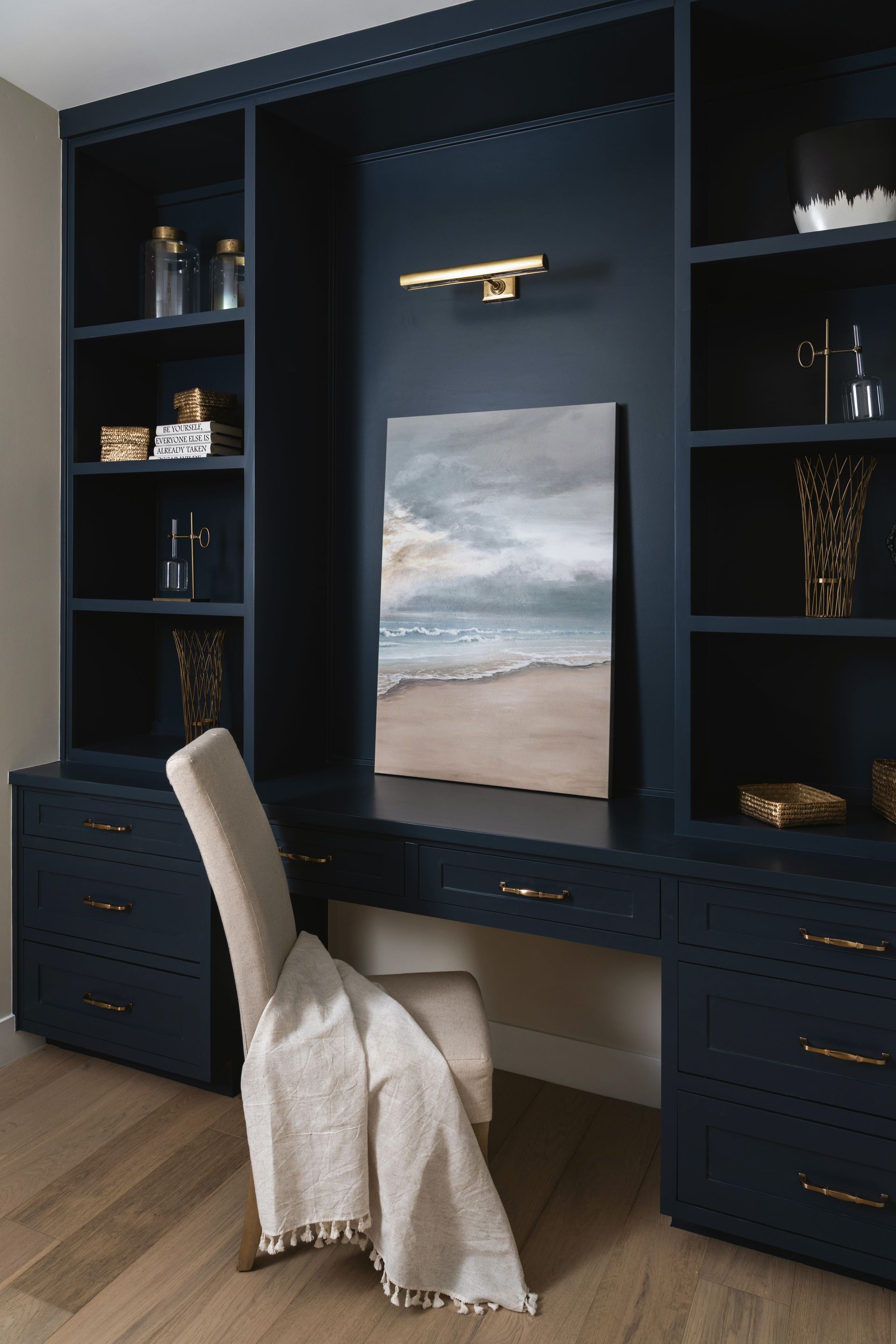 Built-in navy desk with shelving, drawers, and a chair, centered on a landscape painting in a home office.