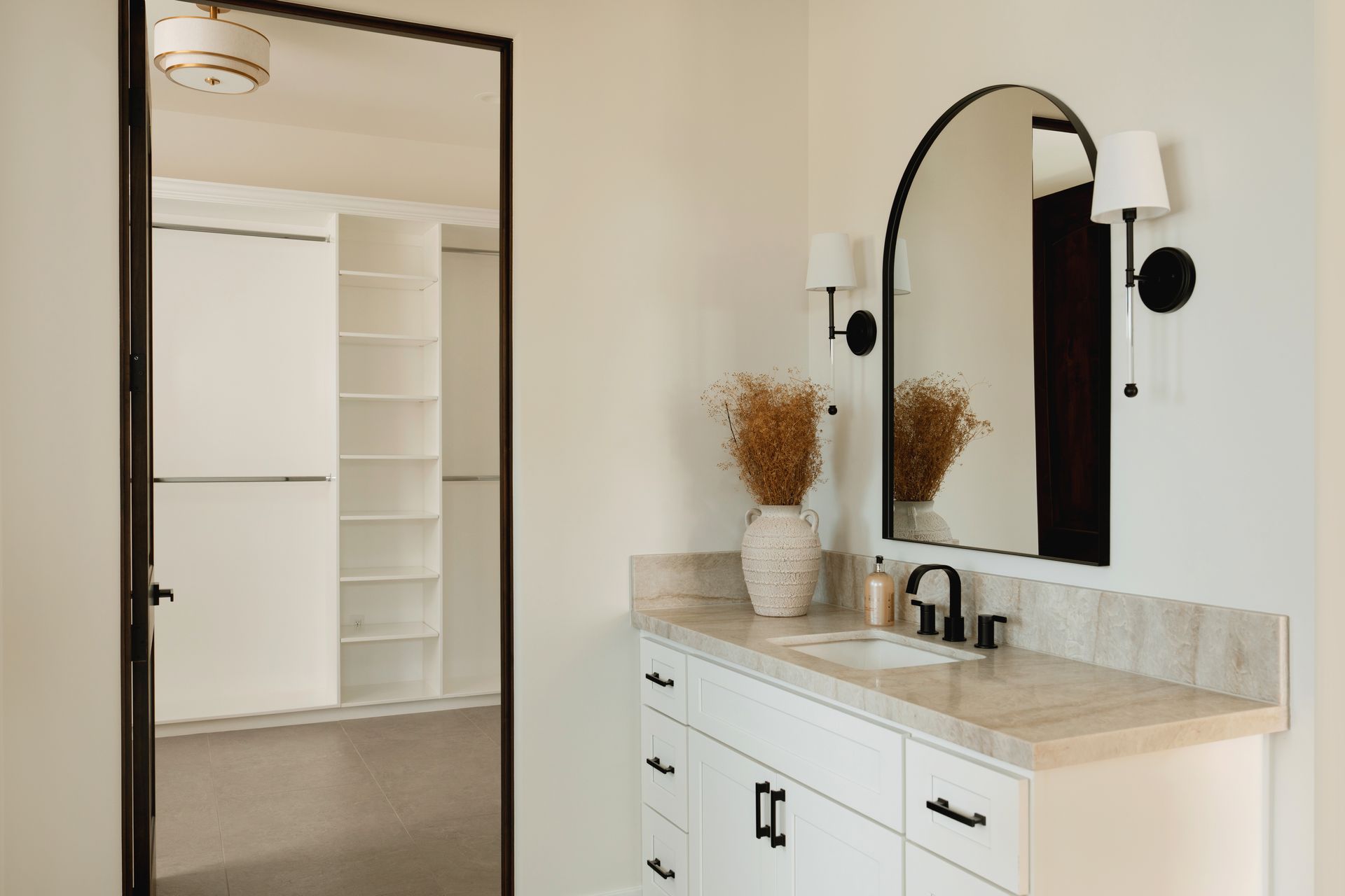 Modern bathroom vanity with arched mirror, white cabinets, and a doorway to a bright closet area
