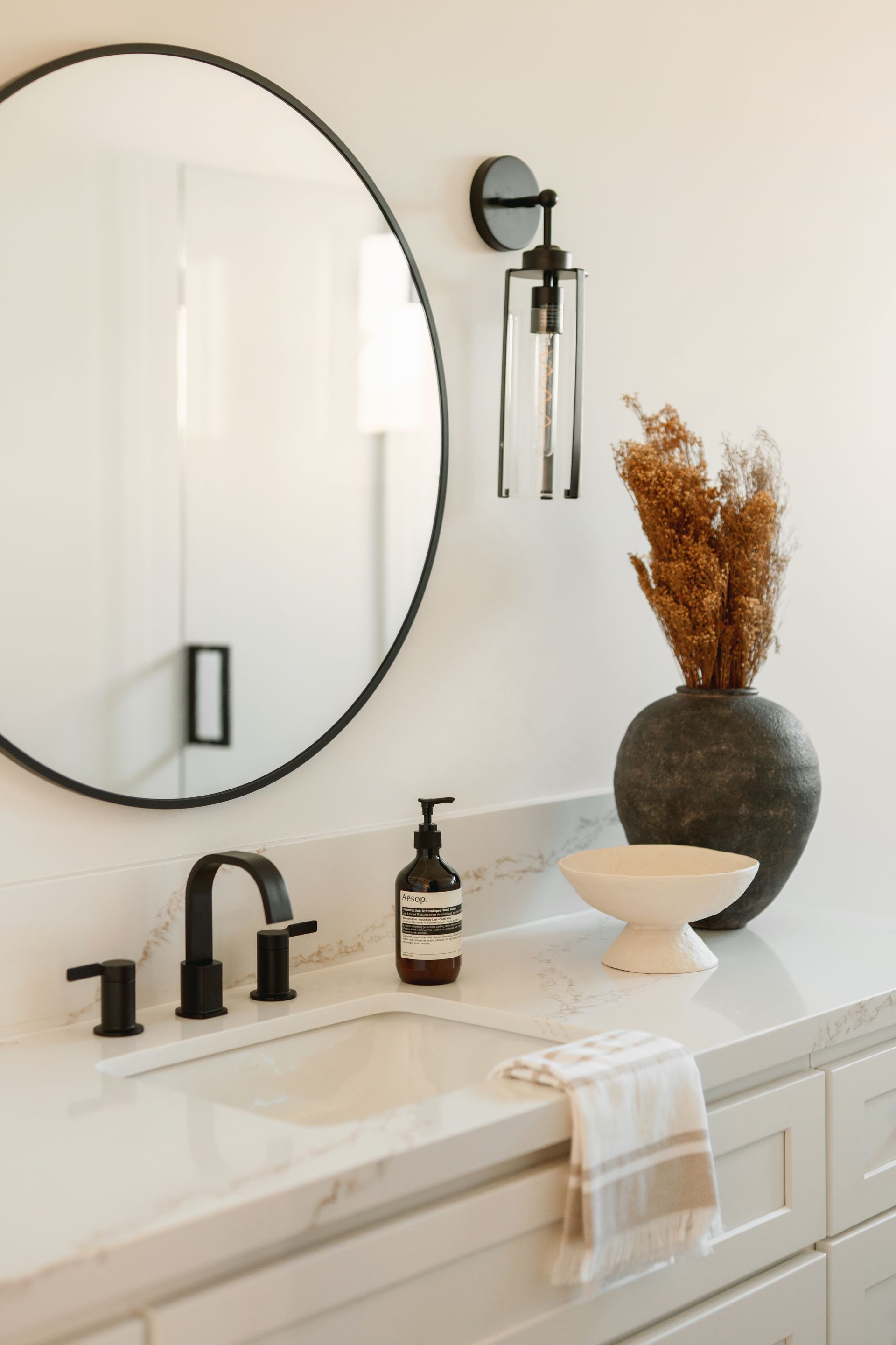 Modern bathroom vanity with round mirror, black fixtures, white countertop, and dried floral decor.