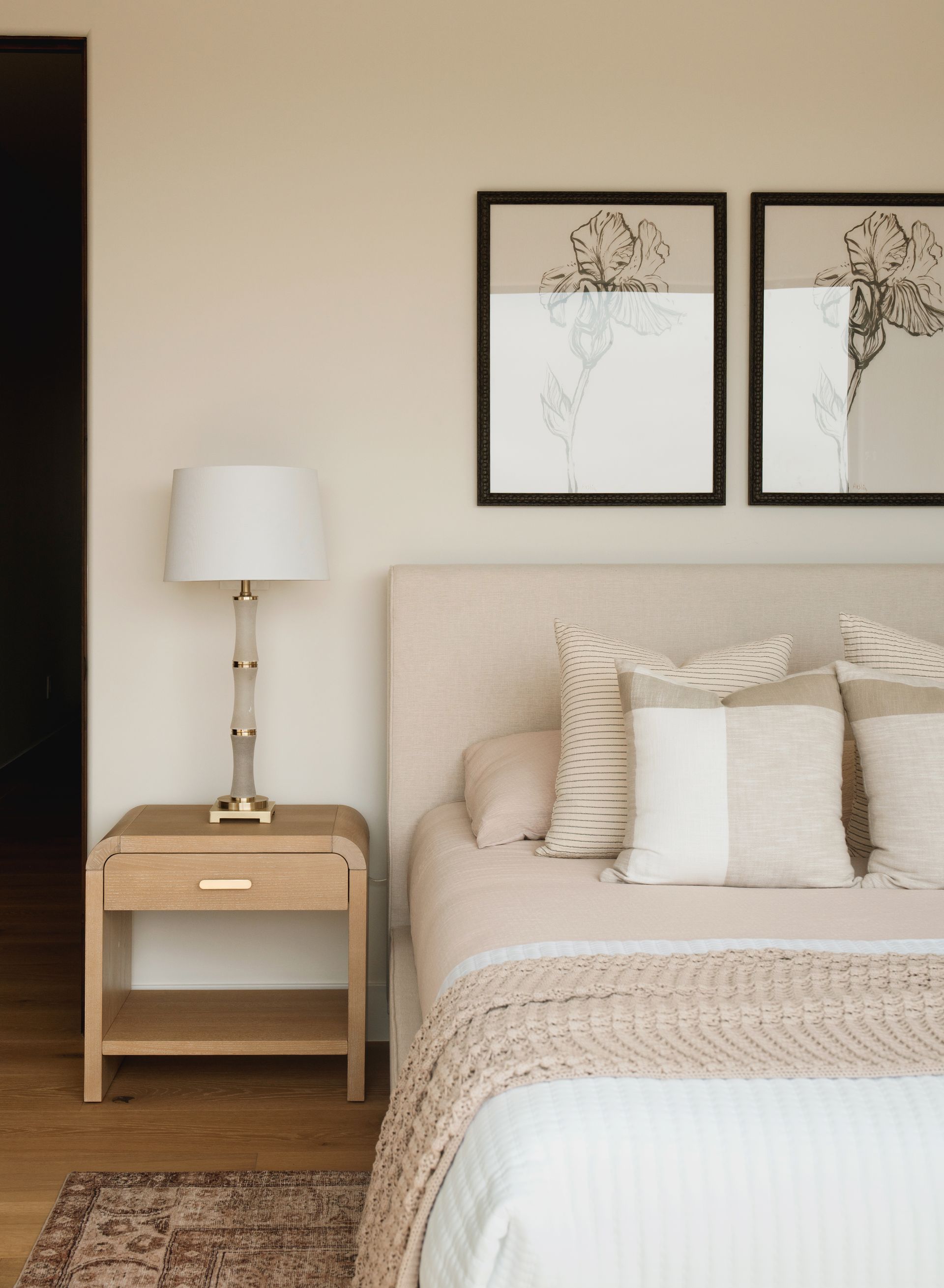 Neutral bedroom with beige bed, wooden nightstand, white lamp, and framed wall art