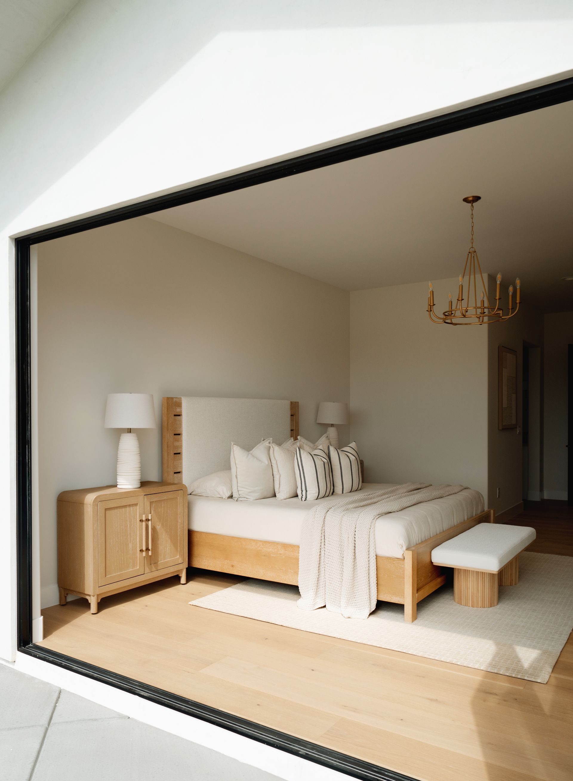 Bright bedroom with a wooden bed, matching nightstands, white linens, and a bench beneath a chandelier.