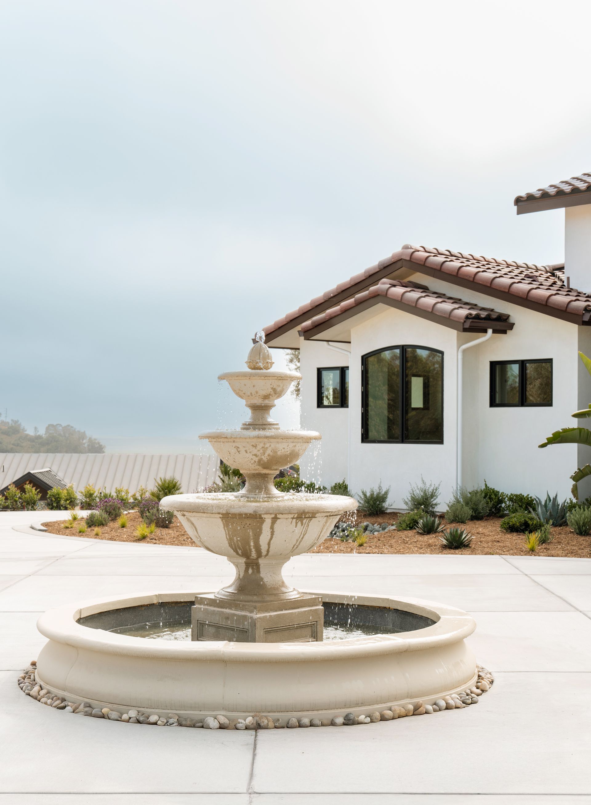 Stone tiered fountain in a landscaped courtyard beside a white house with a tiled roof.
