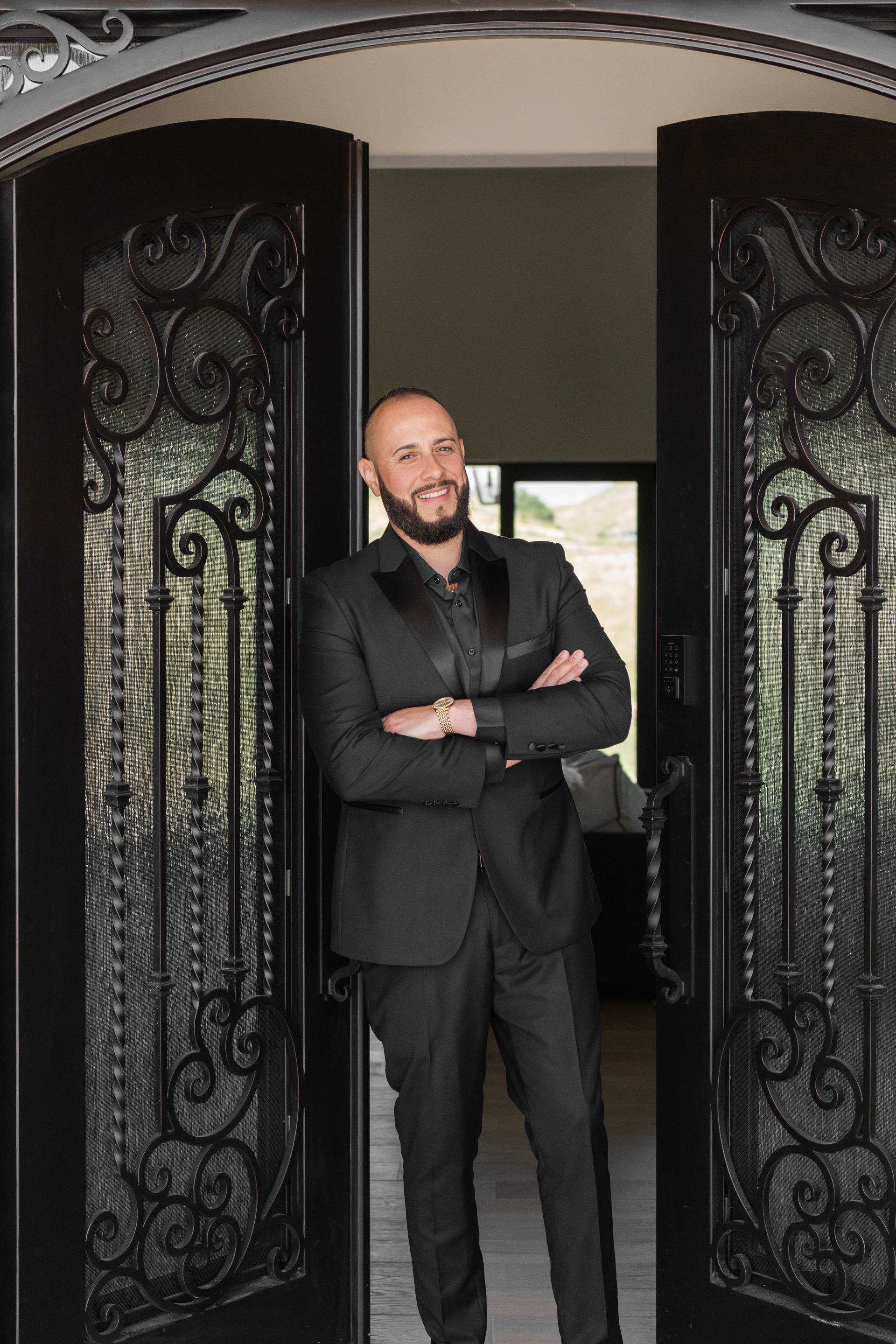 Man in a black suit standing with arms crossed in an ornate doorway